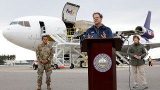 New Hampshire Gov. Chris Sununu, center, speaks to reporters as N.H. National Guard Major General David Mikolaities, left, and Sen. Jeanne Shaheen, D-N.H., right, look on, April 12, 2020, at Manchester-Boston Regional Airport, in Manchester, N.H.
