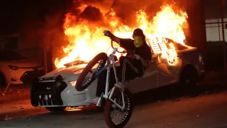 A protester on a bicycle rides past a burning police car during a demonstration next to the city of Miami Police Department, Saturday, May 30, 2020, downtown in Miami.