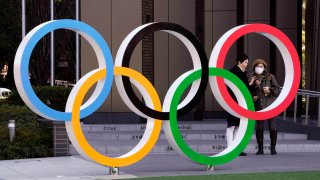 In this Feb. 27, 2020, file photo, visitors wearing face masks stand next to the Olympic Rings monument in front of the Japan Olympic Committee headquarters near the new National Stadium, venue of the Opening and Closing Ceremony of the Tokyo Olympic Games, in Tokyo, Japan.