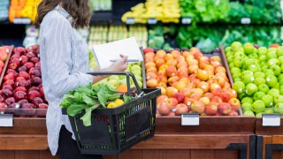 Young woman carries a shopping basket filled with fresh produce. She is shopping for fresh fruit and vegetables in a grocery store.