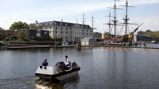 An electric boat steers close to a full-size replica of the 18th century three-mast trading ship Amsterdam at the National Maritime Museum, in Amsterdam, Thursday, May 20, 2021. Already steeped in maritime history, the city's more than 100 kilometers (60 miles) of waterways are to start hosting prototypes of futuristic boats — small, fully-autonomous electric vessels — to carry out tasks including transporting passengers and picking up garbage.