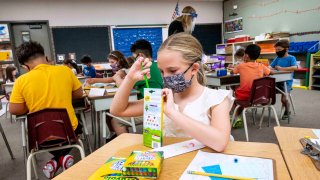 A girl puts a color pencil into its box as she wears a face mask and sits in a classroom.