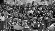 Troops passing in review at Boston's Samuel Adams statue during Queen Elizabeth II's visit on July 11, 1976. Protesters outside Boston City Hall hold up signs in the background.