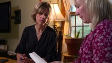 Two woman sit in a room looking at a letter one woman is holding