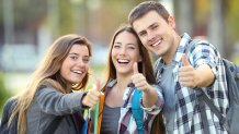 A stock image, captioned, "Three happy students looking at you with thumbs up in an university campus,"