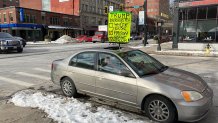 A sign that reads, "Trump belongs in not the White House but prison and an "insane asylum!" being held from the sunroof of a silver sedan.