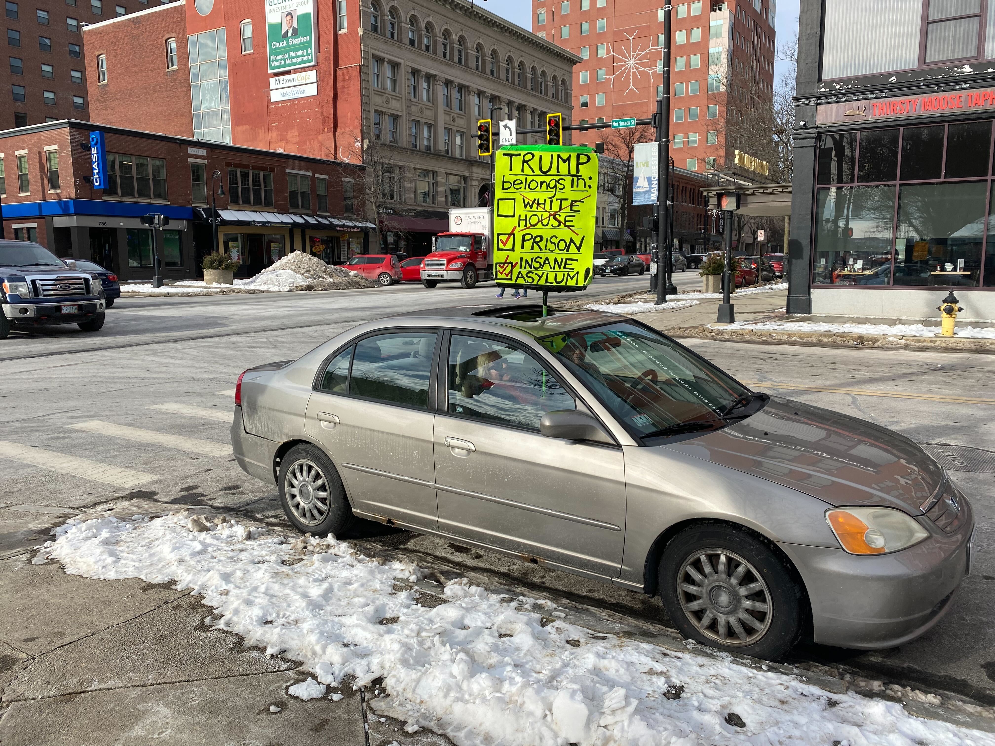 A sign that reads, "Trump belongs in not the White House but prison and an "insane asylum!" being held from the sunroof of a silver sedan.