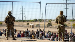 FILE - Migrants wait in line adjacent to the border fence under the watch of the Texas National Guard