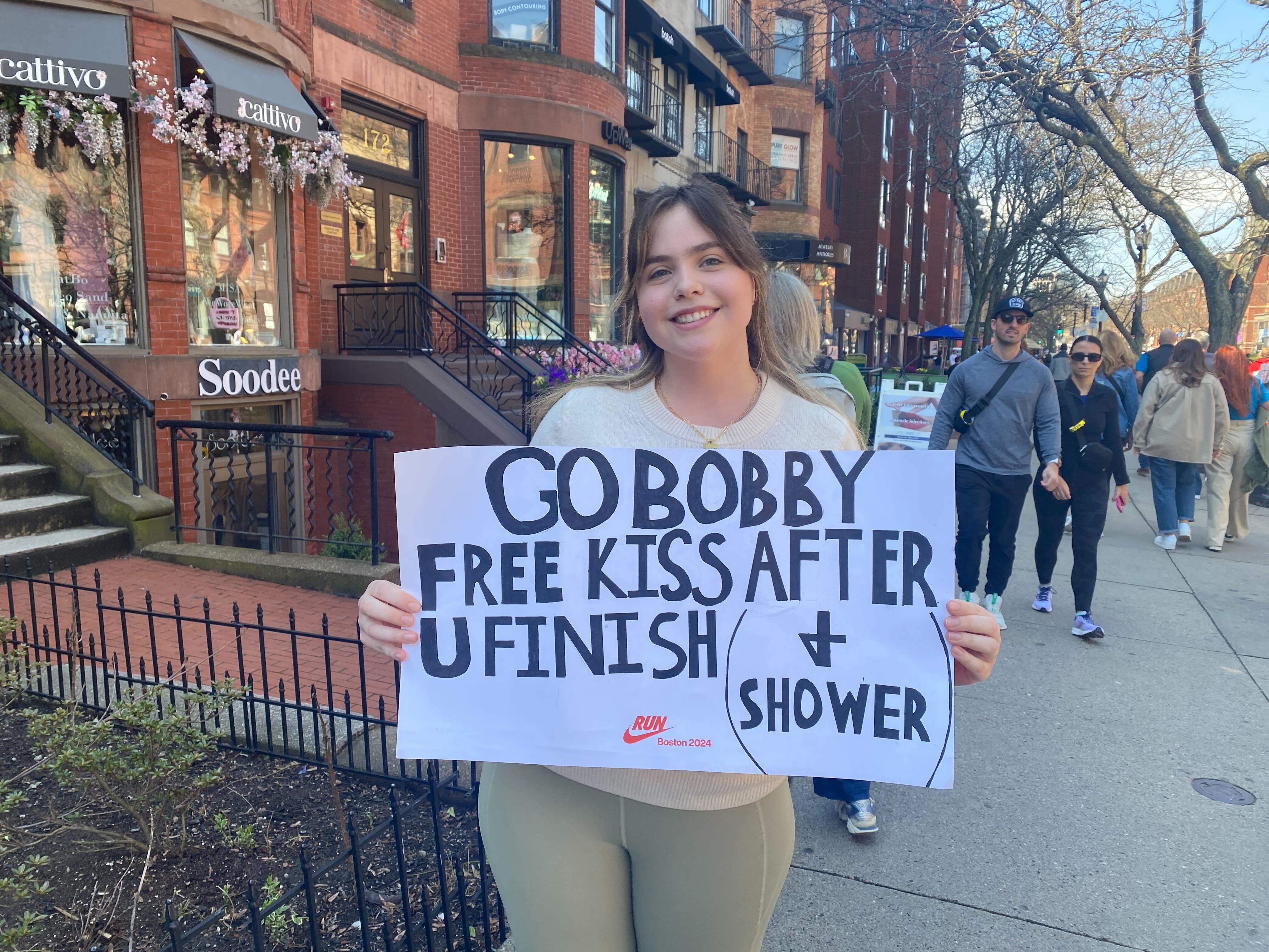A woman on Newbury Street holds a Boston Marathon support sign that reads, "Go Bobby. Free kiss after U finish (+ shower)"