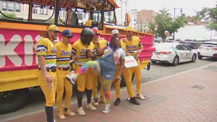 Six Savannah Bananas players posing in front of their Dunkin' and banana themed Duck Boat with a fan.