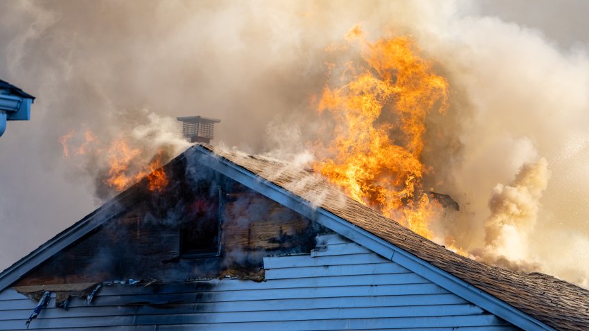 Flames and smoke showing through the room of a white, wooden house with a shingle roof.