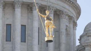 A golden statue of a man holding a spear and a hammer is raised in front of a state house building. The building is light stone with columns and a dome at the top.