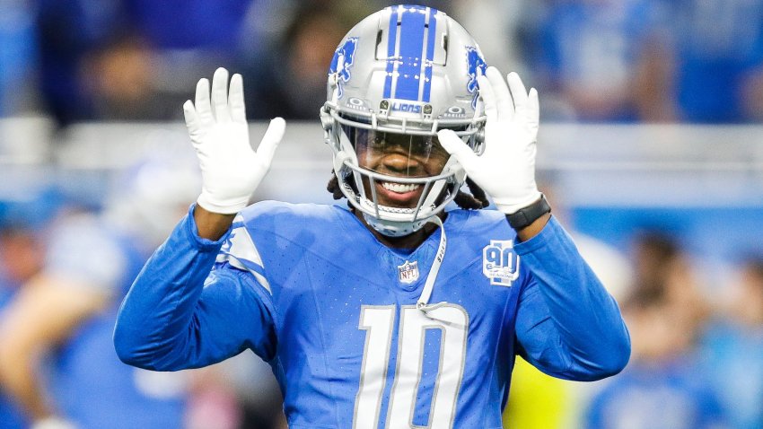 Teddy Bridgewater wears a silver and blue helmet, a light blue Detroit Lions #10 jersey and white gloves. He waves with both hands at the camera and smiles.