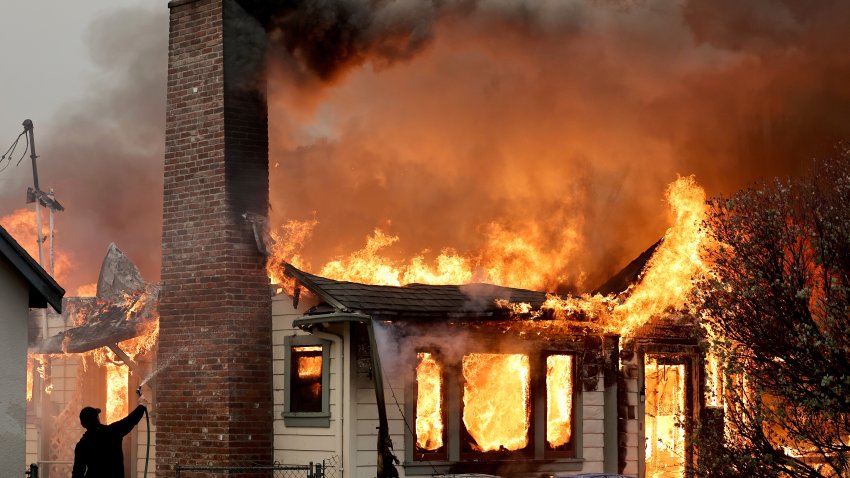 ALTADENA, CALIFORNIA – JANUARY 08: A person uses a garden hose in an effort to save a neighboring home from catching fire during the Eaton Fire on January 8, 2025 in Altadena, California. Over 1,000 structures have burned, with two people dead, in wildfires fueled by intense Santa Ana Winds across L.A. County. (Photo by Mario Tama/Getty Images)