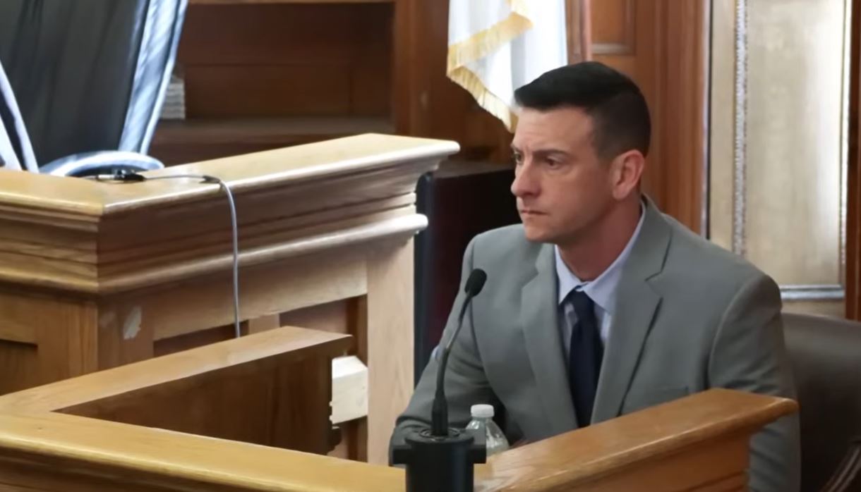 A man wearing a gray suit and a dark tie sits in the witness box in a courtroom
