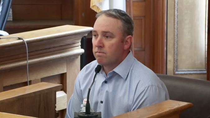 A man wearing a light blue shirt sits in a courtroom witness box