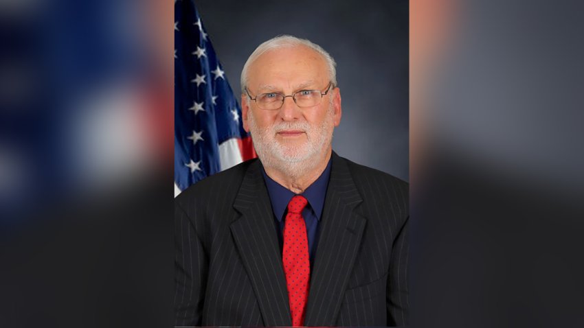 A headshot of Dan Wilson. He has grey hair and a beard and is wearing glasses, a black button-down shirt and a red tie.