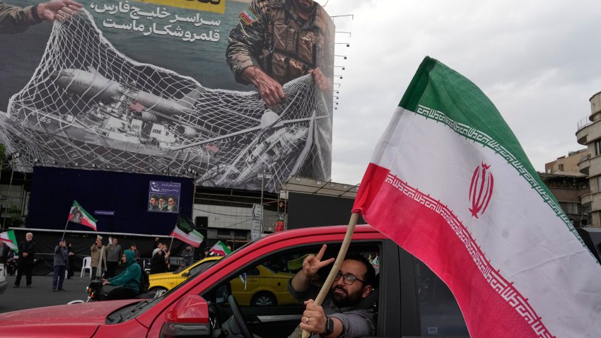 A man flashes a victory sign as he carries an Iranian flag in front of an anti-U.S. billboard depicting the American aircrafts into the Iranian armed forces fishing net with signs that read in Farsi: “The Strait of Hormuz will remain closed, The entire Persian Gulf is our hunting ground,” at the Eqelab-e-Eslami, or Islamic Revolution Square in downtown Tehran, Iran, Monday, April 13, 2026.