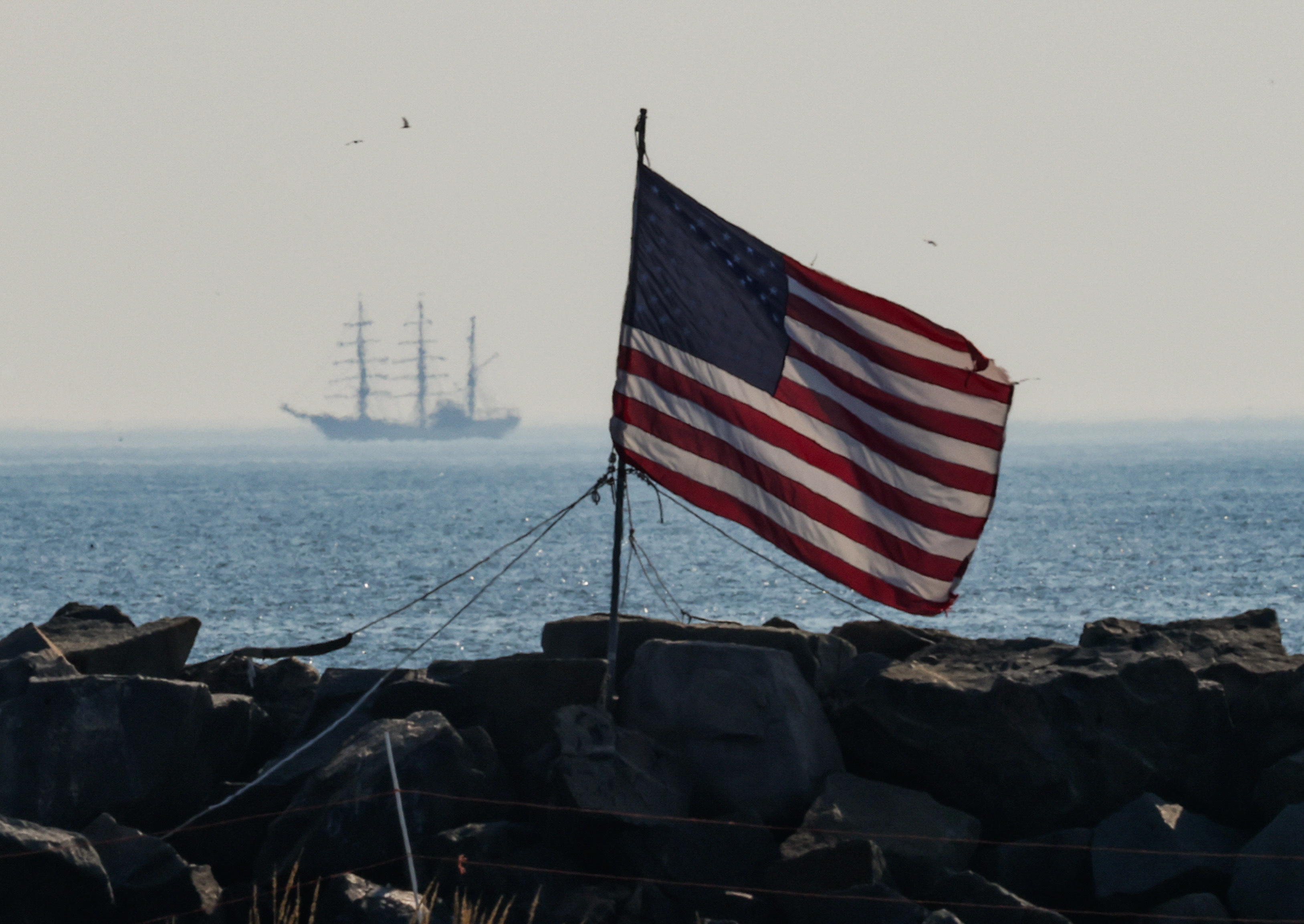 A tall ship is seen in the water off Massachusetts in the distance, behind an American flag in the foreground, ahead of Sail Boston 2026.